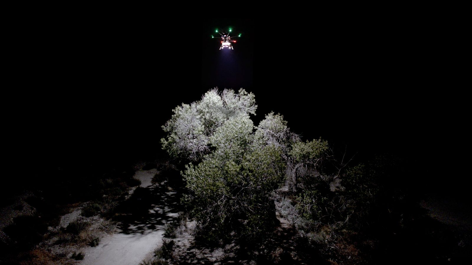 A lighting drone with red and green lights, white beam on a desert shrub