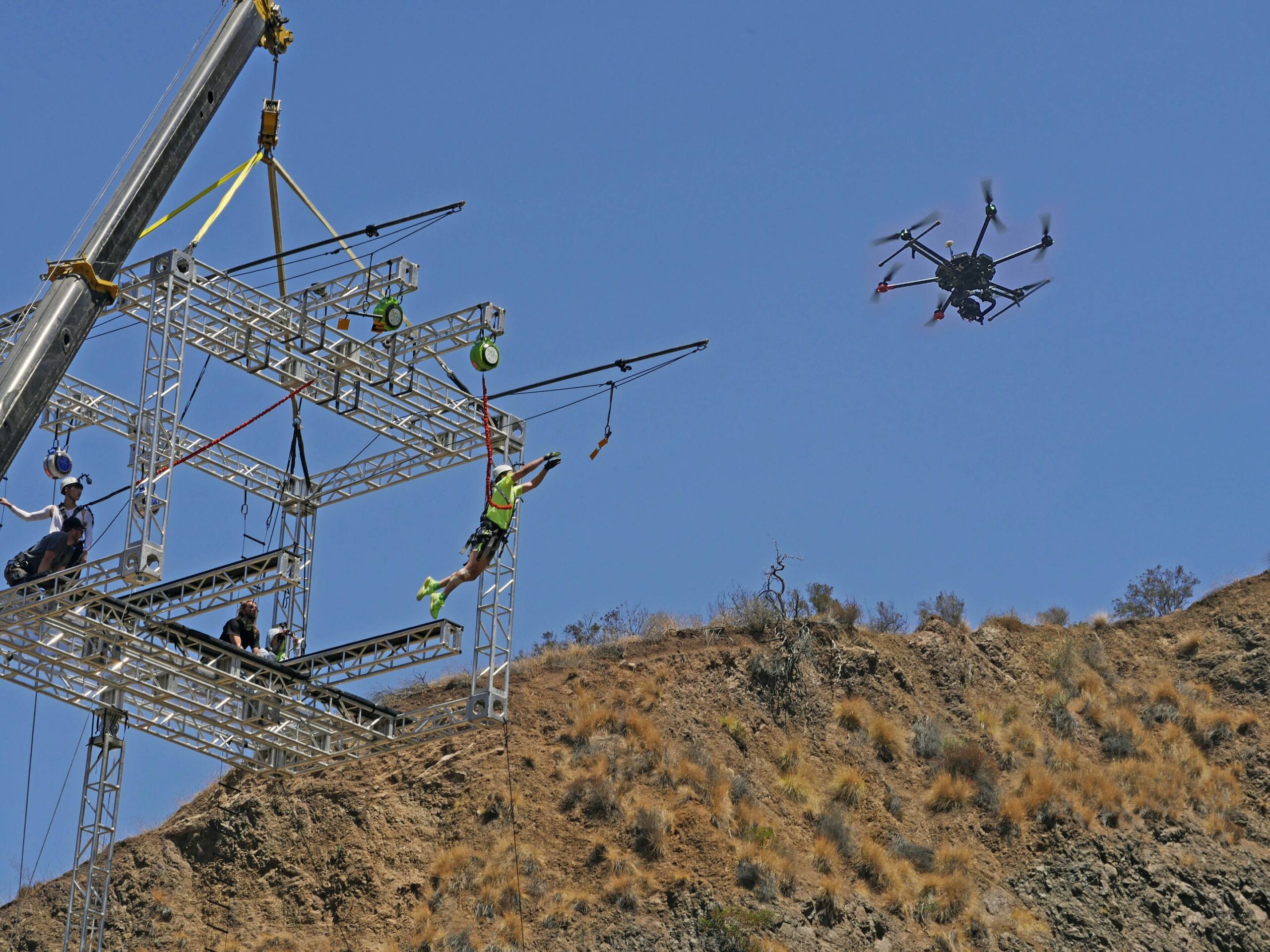 Steel rig on a hillside, camera drone beside it, two people hanging in harnesses