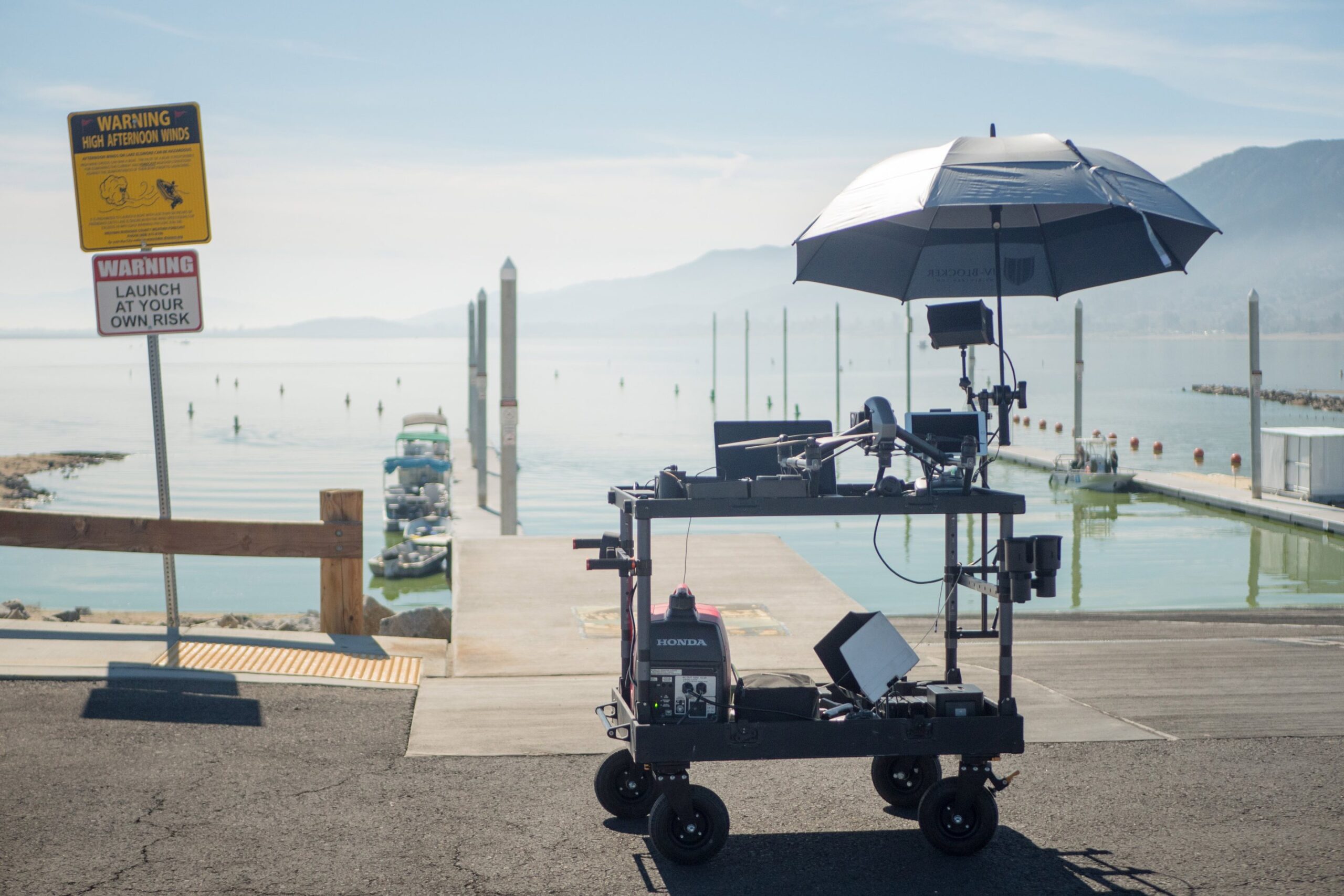Camera drone and monitors under an umbrella on a pier, mountains across the water